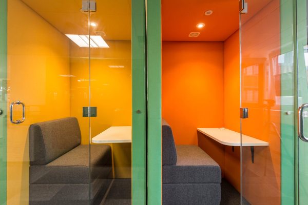 1-36 efurbished enclosed office booths featuring glass doors and vibrant interior color schemes—one in yellow and the other in orange. Each booth includes a grey cushioned bench and a white table, designed for focused work or private meetings. A green partition wall separates the units, contributing to a dynamic and modern workspace. The refurbishment enhances acoustic privacy, visual appeal, and functional versatility in a contemporary office environment.