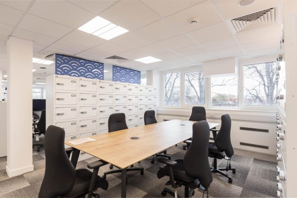 1-42 Refurbished modern office space featuring a large wooden meeting table surrounded by six black ergonomic chairs, complemented by rows of white filing cabinets with labeled drawers for organized storage. Large windows provide ample natural light, while recessed ceiling lighting ensures consistent illumination. The refurbishment enhances workspace functionality, visual clarity, and professional ambiance in a contemporary office setting.