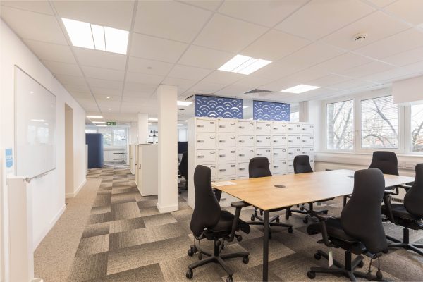 1-44 Refurbished modern office space featuring a large wooden meeting table surrounded by black ergonomic chairs, complemented by white multi-drawer storage cabinets and a blue decorative panel. Large windows provide ample natural light, while gray carpet tiles and recessed ceiling lighting enhance comfort and visibility. A wall-mounted whiteboard adds functionality, reflecting a thoughtful refurbishment focused on collaboration, organization, and contemporary design.