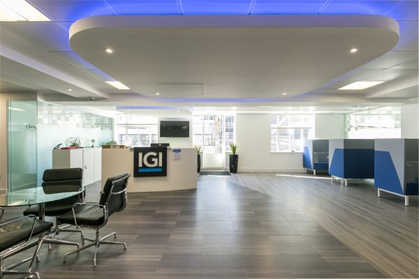 Modern office reception area featuring a sleek desk with the "IGI" logo, black chairs around a glass table, blue and white workstations along the side, dark wood-look tile flooring, large windows with natural light, and decorative plants throughout the space.