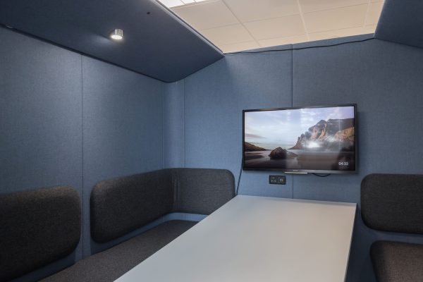 1-60 Refurbished enclosed meeting pod featuring blue fabric-covered acoustic walls and ceiling, a central rectangular table, and gray cushioned bench seating on three sides. A wall-mounted flat-screen television displays a beach scene with cliffs and digital time. The refurbishment enhances privacy, sound insulation, and collaborative functionality in a modern office environment.