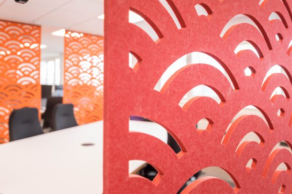 1-67 Refurbished modern office space featuring decorative red and orange wall panels with semi-circular cutout patterns, adding visual interest and acoustic functionality. The open-plan layout includes black ergonomic office chairs and white desks, set in a well-lit environment. The refurbishment enhances workspace aesthetics, promotes collaboration, and improves lighting and spatial efficiency for a contemporary professional setting.
