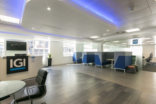 Modern office space featuring multiple workstations with blue and gray partitions, a sleek reception area with a black "IGI" counter, wood-look tile flooring, recessed and blue accent lighting, large windows with natural light, potted plants, and a glass table with black chairs in the foreground.