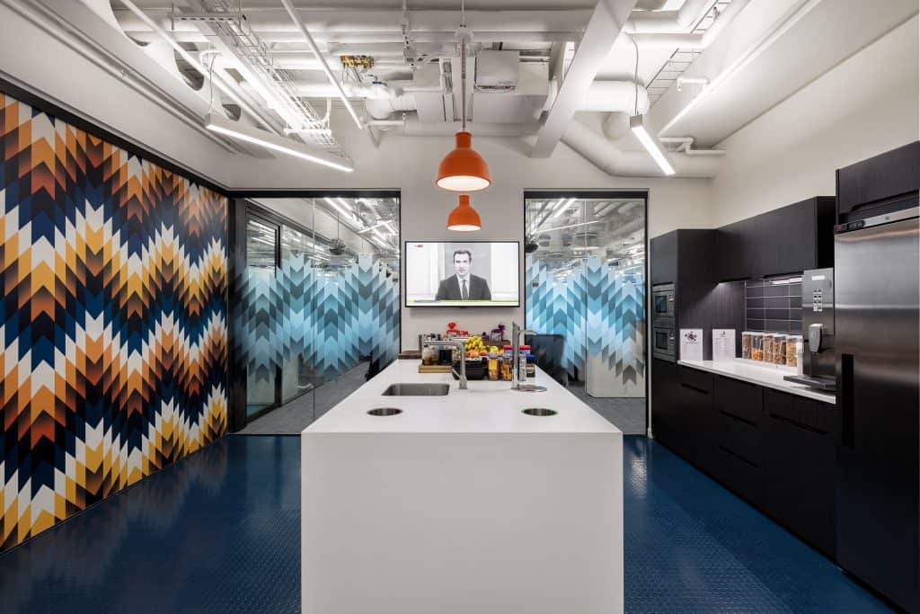 10 - Communal Pantry Modern office kitchen with a central white island featuring two sinks and assorted snacks and drinks. The space includes black cabinets, stainless steel appliances, orange pendant lights, and walls with colorful geometric and blue frosted glass patterns. A screen on the back wall shows a person in business attire on a video call.