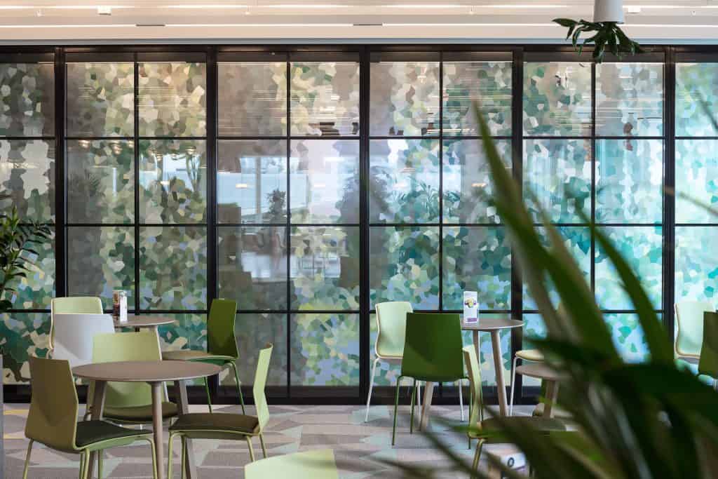 18 - Loose furniture here includes chairs with angled backs in two shades of green from Ocee Design Modern indoor seating area with round tables and green chairs, set against a large glass wall featuring a mosaic pattern in green, blue, and white. Plants are visible in both the foreground and background.