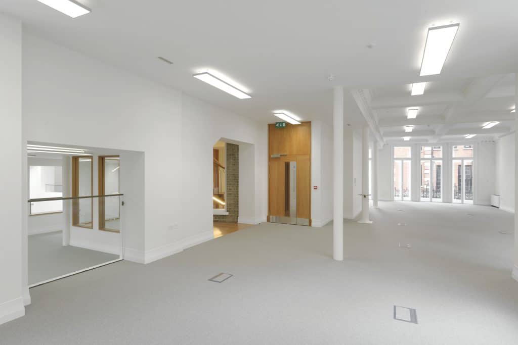 1st Floor_8 Spacious modern interior with white walls and ceiling, light grey carpeted floor, rectangular fluorescent ceiling lights, a central wooden door with a glass panel and exit sign above, open area with glass windows and railing to the left, and large windows at the far end showing buildings outside.