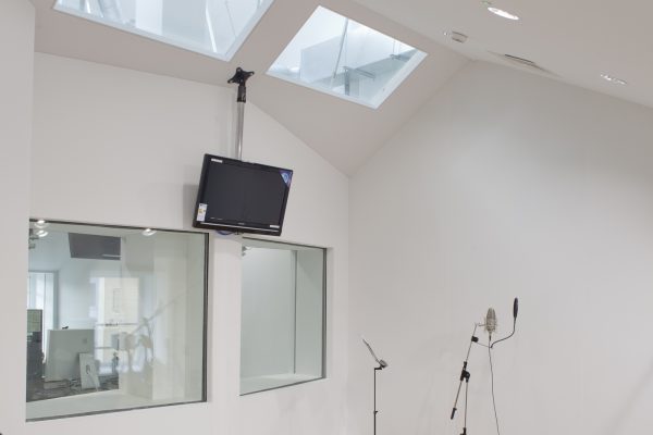 Recording studio featuring two microphones on stands positioned in front of a large interior window that looks into an adjacent room. A wall-mounted television screen is visible to the left, and a skylight in the ceiling allows natural light to illuminate the space.