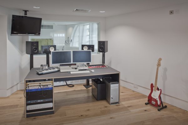 Modern recording studio featuring a desk with dual computer monitors, audio equipment including speakers, a keyboard, and various electronic devices. A red electric guitar stands on a floor stand to the right of the desk. A wall-mounted television is visible on the left, and a window in the background provides a view into an adjacent room. The studio has white walls and a wooden floor.