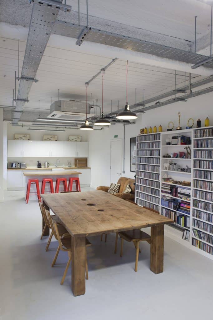 20111117_soundtree_0046 Modern, well-lit room combining a kitchen and living area. The kitchen features white cabinets, a wooden countertop, and three red bar stools, with appliances like a toaster, kettle, and microwave on the counter. Above the cabinets are three framed pictures of crossed arrows. The living area includes a brown leather sofa with patterned cushions, and wall-mounted shelves filled with CDs or DVDs, trophies, books, and small sculptures. A large wooden table sits in the foreground beneath hanging industrial-style lights