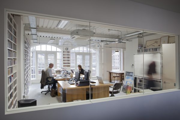 Office space with two individuals seated at desks working on computers, surrounded by shelves filled with books or files. The room features large windows allowing natural light, and various office equipment including telephones and monitors. A third person is walking past in the background.
