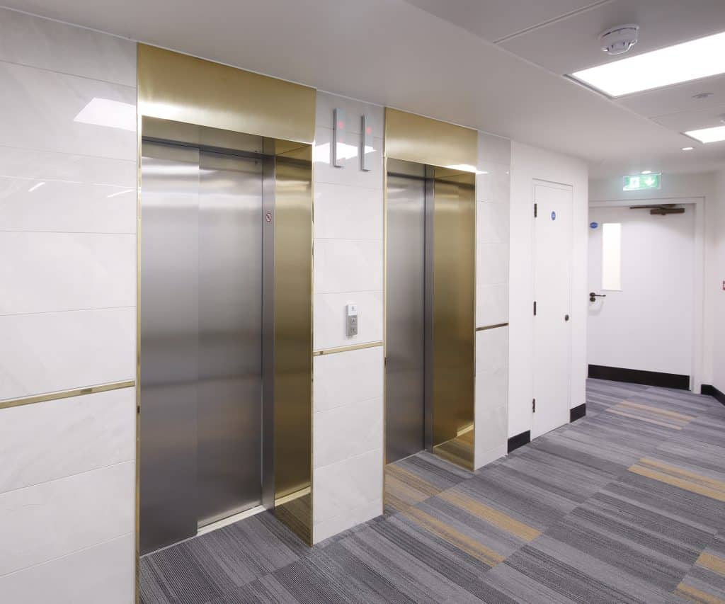 _B4A0508 Hallway featuring gold-framed elevator doors, striped carpeting, and updated wall finishes with gold accents, combining safety enhancements and modern design for a polished, professional environment.