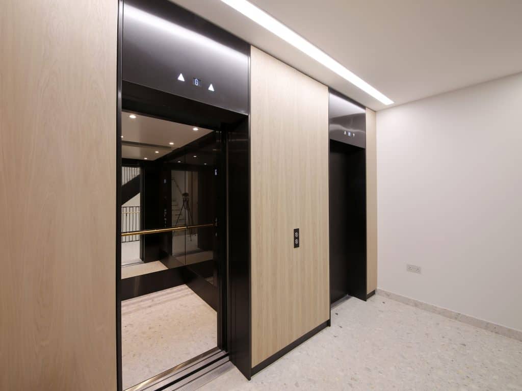 _B4A6536 Modern elevator lobby featuring two black reflective elevator doors set in light wood-paneled walls, with a light-colored marble or tile floor; one elevator is open, revealing a mirrored interior with a handrail and upward-pointing indicator lights above each door.