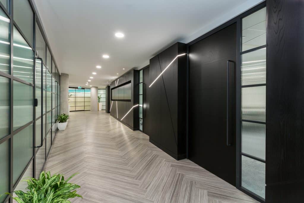 Lift Installation Modern hallway with herringbone-patterned flooring, glass panels on one side, and dark wooden panels with embedded linear LED lights on the other. Recessed ceiling lights and strategically placed plants enhance the sleek, contemporary design.