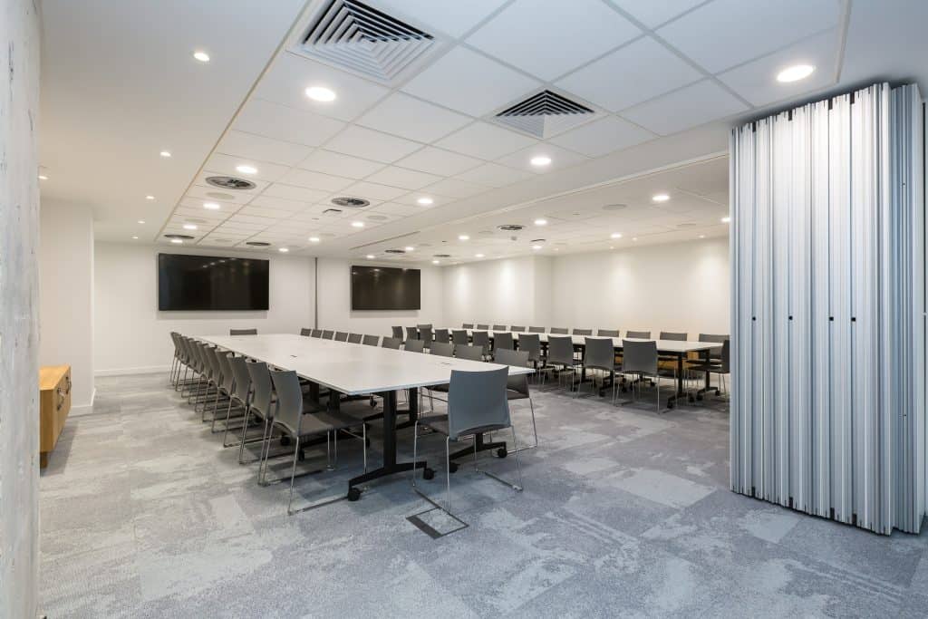 BCS-36 Refurbished conference room featuring two large rectangular tables surrounded by gray chairs, recessed ceiling lights, ventilation units, two wall-mounted flat-screen TVs, light grey carpet tiles, and a folding partition wall.