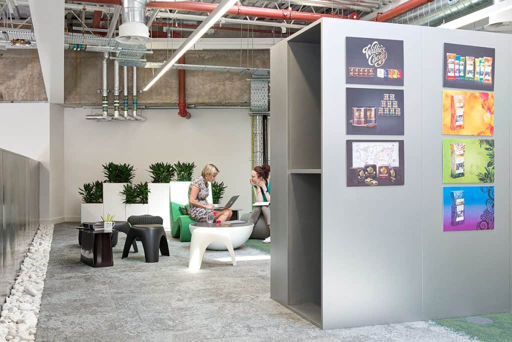 BrandOpus 14 Refurbished office lounge featuring a mix of modern seating, white plant stands, and updated wall displays showcasing product packaging. Exposed ceiling ducts and refreshed décor create a vibrant, collaborative atmosphere.