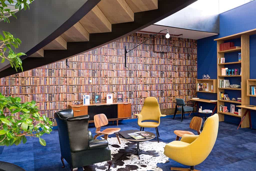BrandOpus 19 Refurbished library and reading area featuring a full-wall bookshelf, vibrant blue carpeting, and a mix of yellow, black, and wooden seating. Added greenery and updated lighting enhance the cozy, modern ambiance, with a staircase hinting at expanded access.