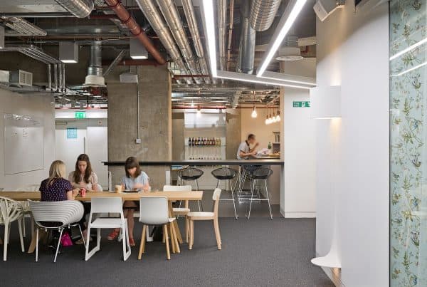 Refurbished office break room featuring a wooden communal table with mixed seating, a modern bar-style counter with bottle display, and exposed ceiling pipes. Updated lighting and concrete-accented white walls create a refreshed, social-friendly space.
