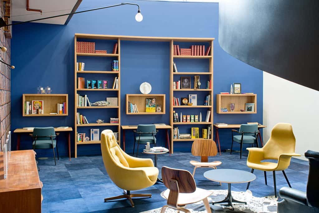 BrandOpus 27 Refurbished reading and study area featuring built-in wooden shelving, updated blue walls, and modern desks with overhead storage. Casual seating with stylish chairs and tables enhances the refreshed, contemporary ambiance ideal for focused work or relaxation.
