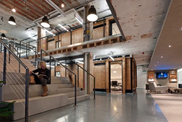 Modern industrial-style office space with exposed ceilings and ductwork, featuring wooden walls and beams. A person is seated on a staircase on the left side, reading. The open layout includes visible meeting rooms in the background, lounge areas with sofas and chairs on the right, and a wall-mounted television. Lighting consists of hanging lamps and recessed fixtures.
