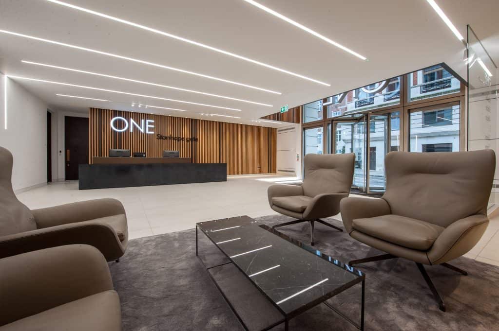 DSC_3141-HDR Modern office lobby with a reception desk set against a wall of vertical wooden slats, illuminated signage reading "ONE Stanhope Gate," two beige armchairs and a glass coffee table on a dark rug in the foreground, and large windows allowing natural light to fill the space.