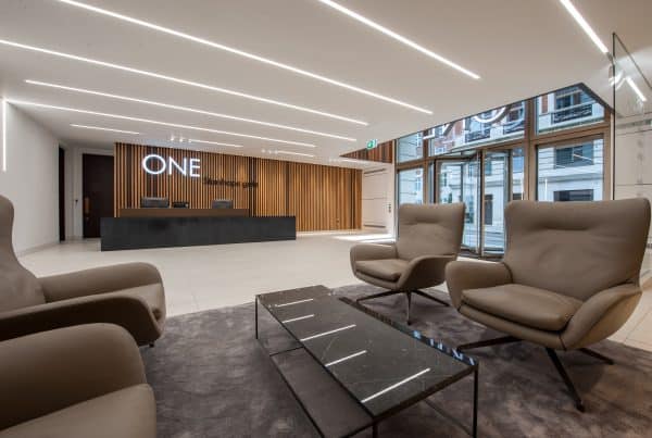 Modern office lobby with a reception desk set against a wall of vertical wooden slats, illuminated signage reading "ONE Stanhope Gate," two beige armchairs and a glass coffee table on a dark rug in the foreground, and large windows allowing natural light to fill the space.