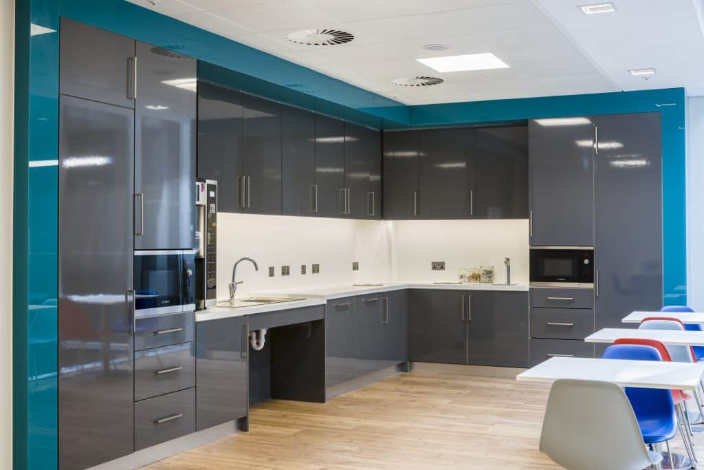 GGARDNER-7291 Modern kitchen with glossy gray cabinets, white countertops, and blue accents. Features include a sink, built-in oven and microwave, electrical outlets on the backsplash, and wooden flooring. Colorful chairs surround tables in the foreground.