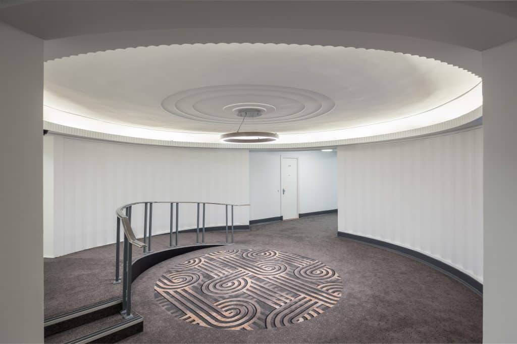 IMG_0010 A stylish, circular hallway featuring a curved railing and a decorative ceiling with circular patterns. Soft lighting creates a warm ambiance, and the floor is covered with plush carpet displaying a geometric design. A door is visible in the background, leading to another room.