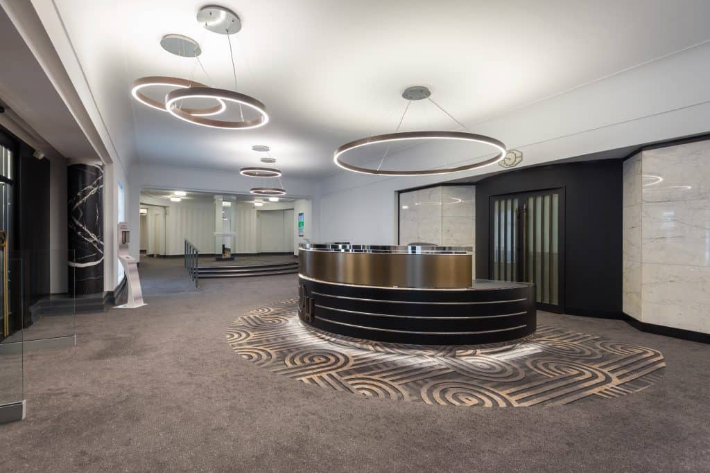 IMG_0037 A newly refurbished lobby or reception area showcasing a contemporary and high-end design. At the center is a circular reception desk with a sleek black and metallic finish, serving as the visual anchor of the space. Suspended above the desk are large, modern circular light fixtures that provide both ambient lighting and a striking design element. The flooring around the desk features an intricate, custom pattern that adds texture and visual interest, suggesting a premium material choice. In the background, a staircase leads to an upper level, indicating a multi-floor layout. Additional ceiling-mounted lighting fixtures contribute to a well-lit, welcoming atmosphere. The refurbishment emphasizes luxury, spatial flow, and a strong first impression for visitors.