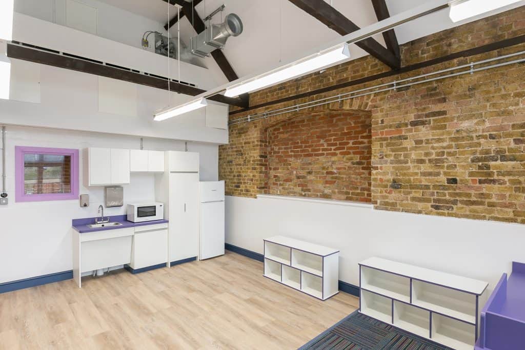IMG_5185 Refurbished modern kitchen area featuring white cabinetry, integrated refrigerator and microwave, and vibrant purple countertops. The space includes white open-shelf storage units with purple trim, set against a textured brick wall for added character. Light wood flooring and exposed ceiling beams with fluorescent lighting contribute to a contemporary industrial aesthetic. The refurbishment enhances functionality, color contrast, and spatial efficiency in a stylish, updated interior.