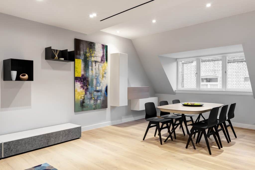 JamesSt-04 Refurbished modern dining room featuring a rectangular dining table surrounded by six black chairs, set against white walls and light wooden flooring. Decorative black wall shelves hold a vase and incense sticks, while a long bench or cabinet with a grey cushion adds functional seating and storage. An abstract multicolored painting and asymmetrically mounted white cabinets enhance visual interest. Large windows and recessed ceiling lighting contribute to a bright, contemporary atmosphere, reflecting a thoughtful refurbishment focused on style, comfort, and spatial efficiency.