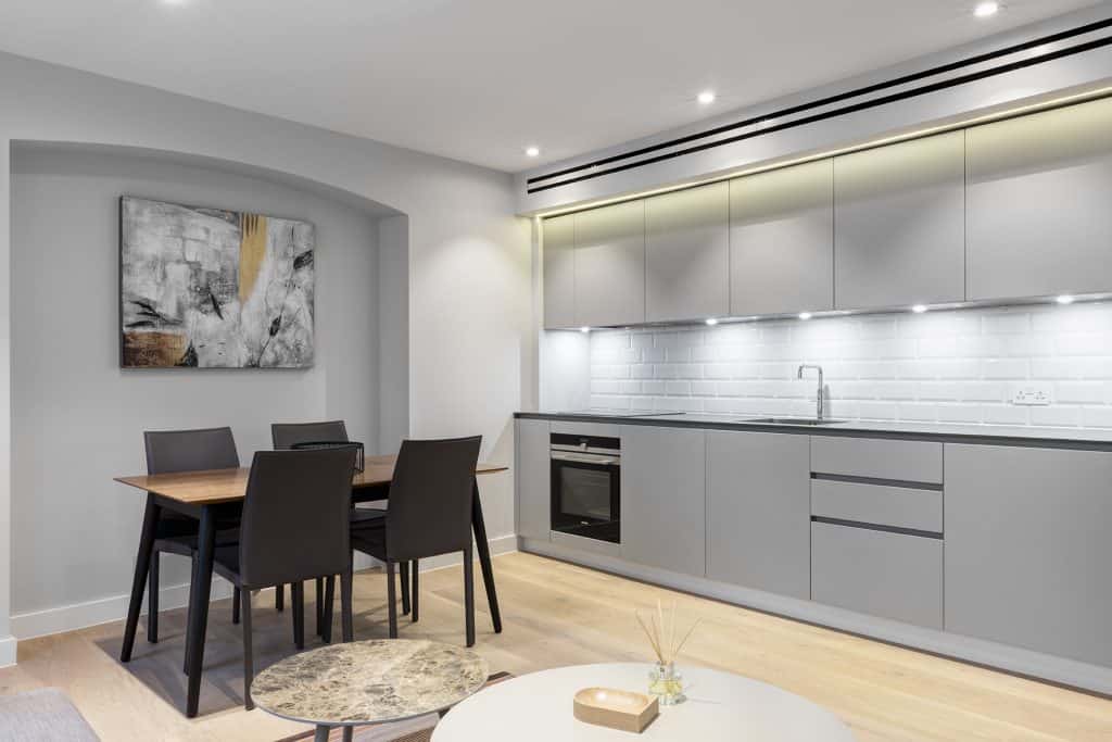 JamesSt-12 Refurbished modern kitchen and dining area featuring sleek grey cabinetry with integrated under-cabinet lighting, built-in oven, and minimalist white subway tile backsplash. A wooden dining table with four dark-toned chairs sits adjacent to the kitchen, complemented by a contemporary abstract painting in grey, black, white, and yellow tones. Light-colored wooden flooring enhances the space’s warmth and cohesion, reflecting a high-end refurbishment focused on functionality, aesthetic harmony, and modern living.