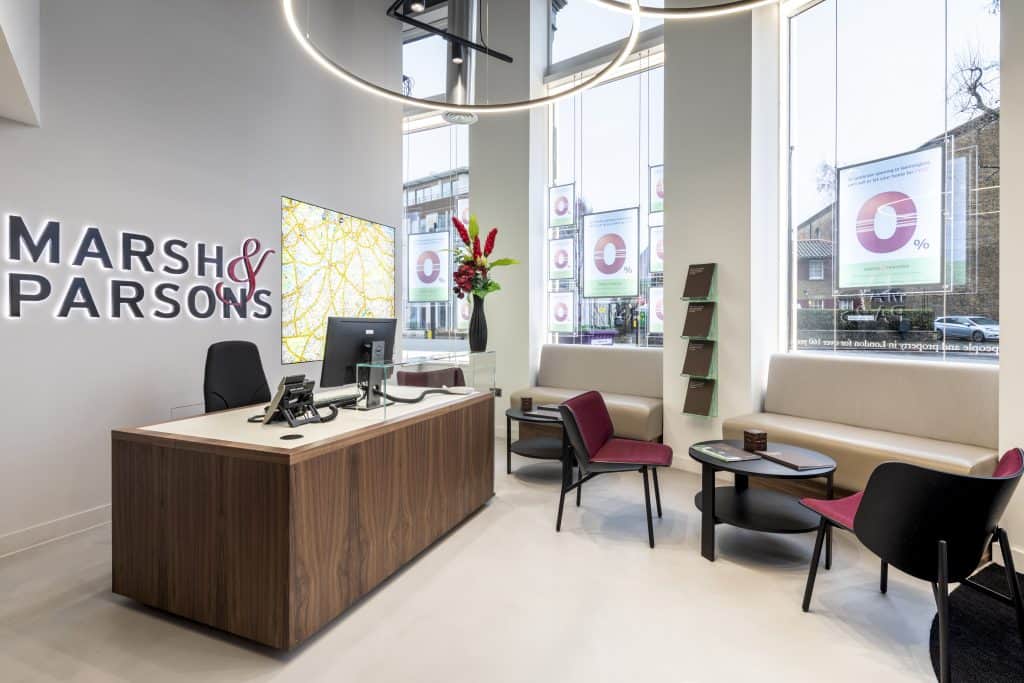 Marsh&Parsons_005 Refurbished Marsh & Parsons reception area featuring a modern wooden desk setup, illuminated branding, and updated seating with beige sofas and vibrant red and black chairs. Enhanced natural light from large windows and refreshed décor, including promotional posters and floral accents, complete the contemporary transformation.
