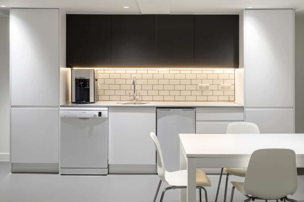 Marsh&Parsons_022 Refurbished office kitchen featuring sleek white cabinetry, updated tiled backsplash, integrated appliances, and contrasting black overhead units. Enhanced lighting and a clean dining setup with white table and chairs reflect a modern, refreshed break area.