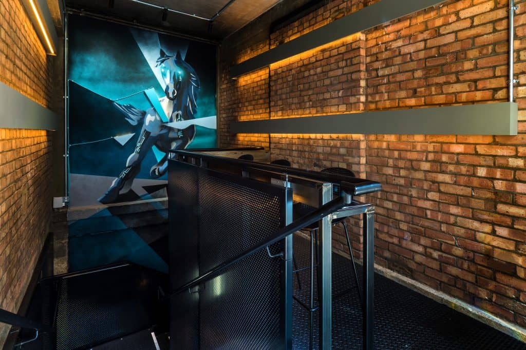 OLYMPUS DIGITAL CAMERA A stylish staircase leading downwards, surrounded by exposed brick walls. A vibrant mural featuring a dynamic figure in blue and black adds a striking focal point to the space. The staircase is made of dark metal, and modern lighting strips along the walls enhance the industrial-chic ambiance.