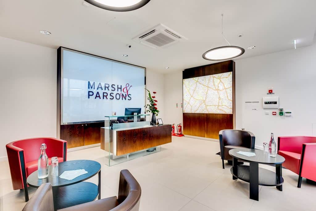 STREATHAM OFFICE-2 Refurbished office reception area of Marsh & Parsons featuring a sleek wooden and glass desk, updated signage, and modernized seating with red and black chairs. A newly installed illuminated map and refreshed décor, including plants and minimalist finishes.