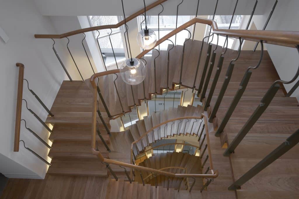Stairs_4th Downward view of a modern spiral staircase with wooden steps, metal railings, and hanging glass pendant lights, creating a repeating geometric pattern.