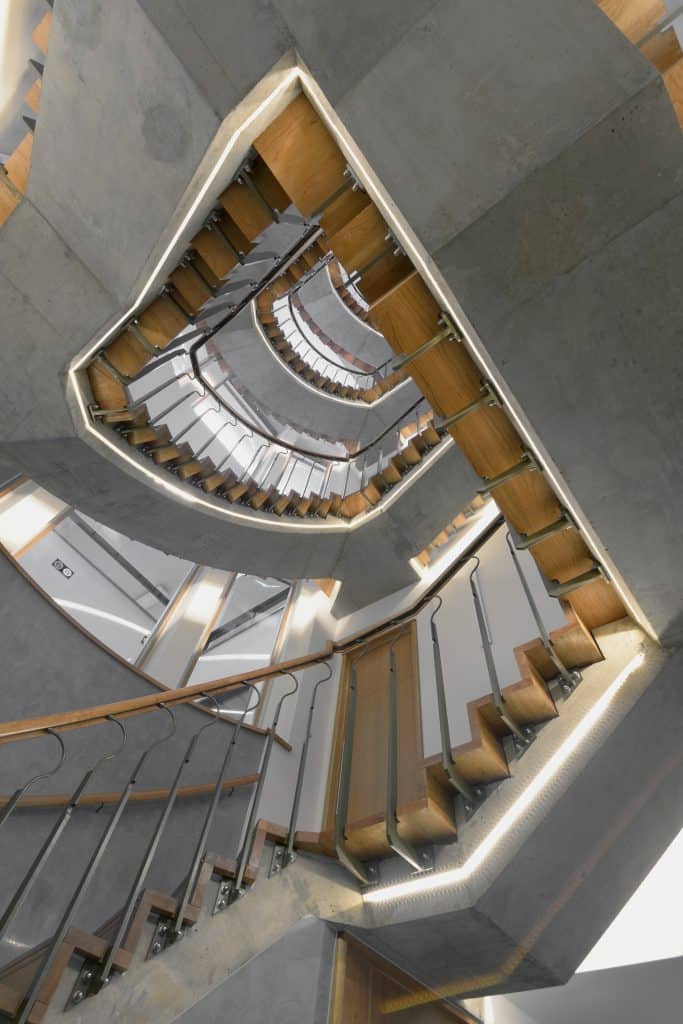 Stairs_looking up Upward view of a modern spiral staircase with wooden steps, metal railings, and concrete walls, creating a geometric pattern with repeating shapes and lines.