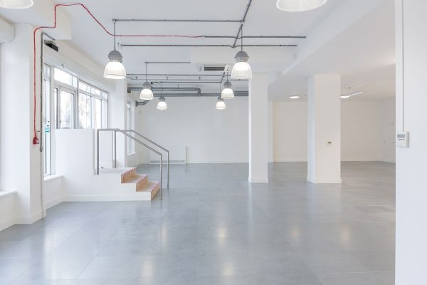 1-10 Refurbished open-plan interior space featuring a modern fit-out with white walls and grey tiled flooring. Large windows on the left side allow natural light to fill the room, enhancing its spacious and versatile layout. Industrial-style pendant lights hang from the ceiling, contributing to a contemporary aesthetic. A small staircase with metal railings leads to an elevated platform, offering potential for zoned usage. The clean, minimal design supports flexible fit-out options for office, studio, or commercial use.