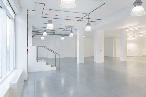 1-12 Refurbished open-plan interior space prepared for fit-out, featuring white walls and a grey floor with a clean, modern finish. Large windows on the left side allow natural light to flood the room, enhancing its spaciousness. Exposed ceiling services, including pipes, ducts, and suspended lighting, reflect an industrial-modern aesthetic. A small staircase with a metal railing leads to an elevated platform, suggesting flexible zoning potential. The space is well-lit and ready for further configuration, ideal for commercial or creative use.