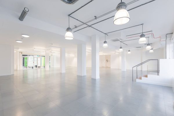 Workspace design london Refurbished commercial interior featuring a spacious, modern fit-out with light grey tiled flooring and crisp white walls. Evenly spaced white columns support the open-plan layout, while industrial-style pendant lighting and exposed ceiling piping contribute to a contemporary aesthetic. A small staircase with a metal railing leads to an elevated platform, offering flexible zoning options. Glass doors at the rear provide access to adjacent areas or exterior space, enhancing natural light and connectivity. The clean, minimal design supports a wide range of future fit-out configurations.