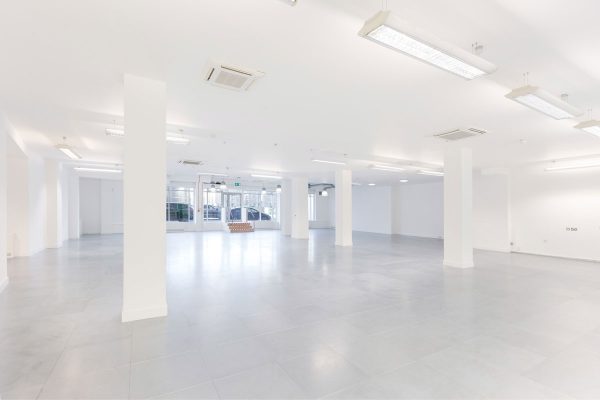 1-8 Refurbished commercial interior featuring a spacious, open-plan layout with white walls and grey flooring. Evenly spaced white columns support the structure, while ceiling-mounted lighting and air conditioning units reflect a modern services fit-out. At the far end, a glass double-door entrance with surrounding windows allows natural light to enter, enhancing visibility and openness. The clean, neutral palette and infrastructure readiness make the space ideal for flexible fit-out configurations, suitable for retail, office, or showroom use.