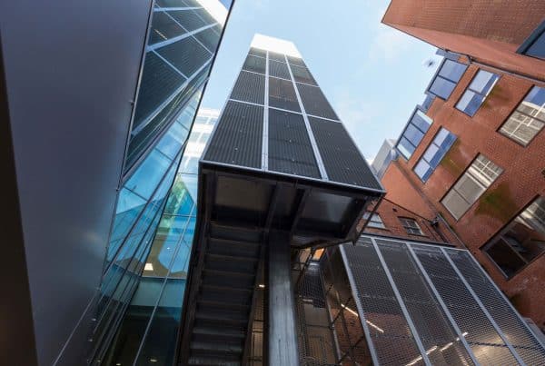 Upward view of a refurbished architectural structure showcasing a modern fit-out with metal paneling and an external staircase. The central building contrasts with surrounding brick-facade structures, highlighting a blend of contemporary and traditional materials. Glass elements and clean lines reflect a design upgrade focused on urban integration and vertical circulation. The visible sky and open angles emphasize the scale and openness of the redevelopment.