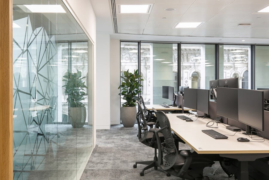 Refurbished modern office interior featuring a high-spec fit out with ergonomic workstations, sleek glass partitioning with geometric designs, and abundant natural light from large windows. The space integrates contemporary furnishings and greenery, highlighting a professional and revitalized working environment.