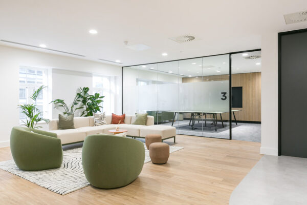 Refurbished office space featuring a stylish fit out with a cozy lounge area furnished with green chairs, a beige sectional sofa, and modern stools on a patterned rug. Adjacent is a glass-enclosed conference room with frosted panels and contemporary furnishings, showcasing a well-executed refurbishment with mixed flooring and integrated greenery.