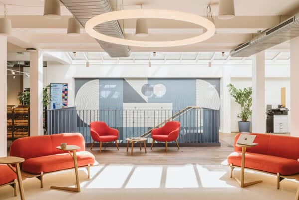 Modern office lounge area with red sofas and armchairs arranged around small wooden tables. A blue wall with geometric patterns and a railing is in the background. The space features large windows, decorative plants, exposed ceiling ductwork, a laptop on one table, and a printer/copier machine on the right.