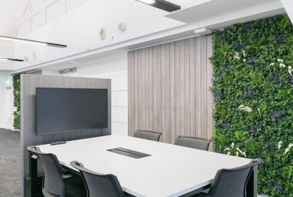 Modern conference room with a large white table surrounded by chairs, a flat-screen TV mounted on a wooden panel, a green plant-covered wall, and recessed lighting with ventilation fixtures on the ceiling.