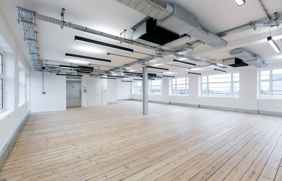Large open plan CAT A office recently completed refurbishment, with wooden flooring, white walls, exposed ductwork and electrical conduits on the ceiling, modern lighting fixtures, multiple large windows on the right letting in natural light, and a door with an emergency exit sign at the far end.