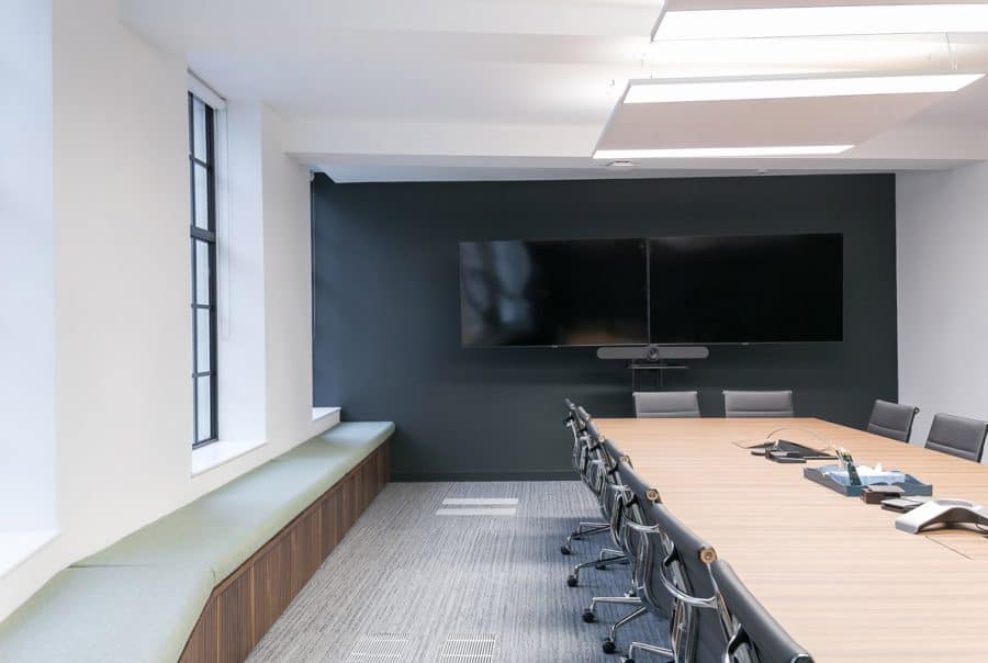 Modern conference room with a long wooden table surrounded by black office chairs, large flat-screen TV mounted on a dark wall, large windows on one side with a built-in bench and green cushions, carpeted floor, and ceiling lights.