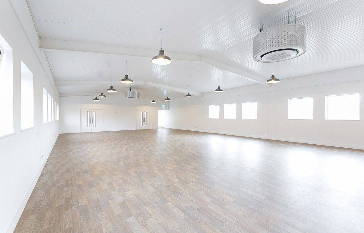 A large, refurbished function room with wooden flooring, white walls and ceiling, multiple windows on the right side letting in natural light, and glass-panel double doors at the far end. Hanging light fixtures provide additional illumination in the spacious, versatile area.