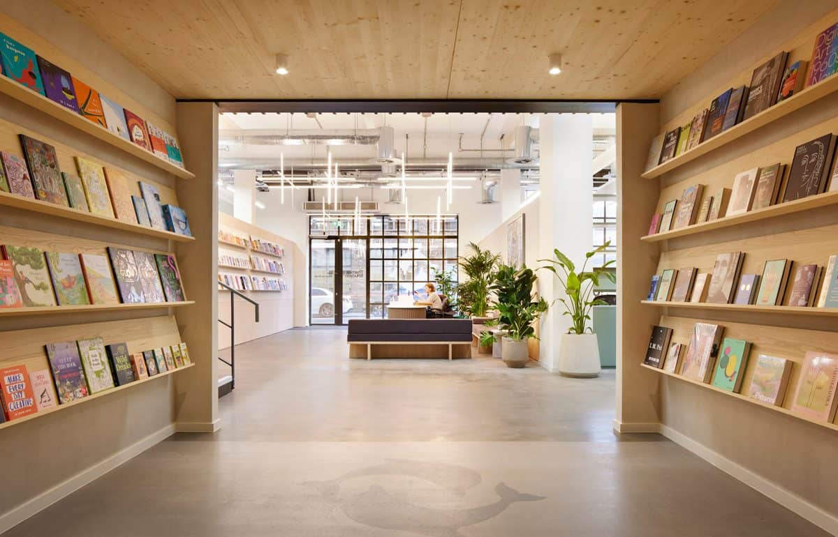 Interior of a modern publishers office with wooden shelves filled with books on both sides. A receptionist sits at a desk in a seating area surrounded by large potted plants. The space is well-lit with hanging contemporary lights and natural light from large windows.